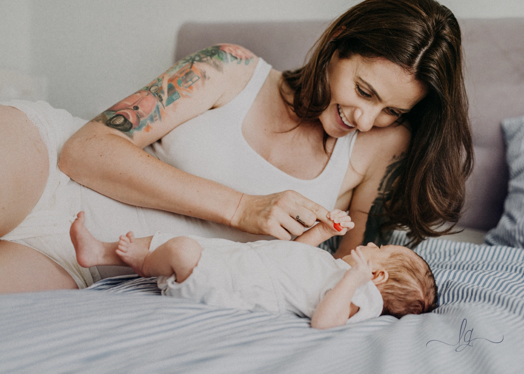A close-up photo of a mother with a tattoo on her arm smiling down at her baby as they lie on a bed together.