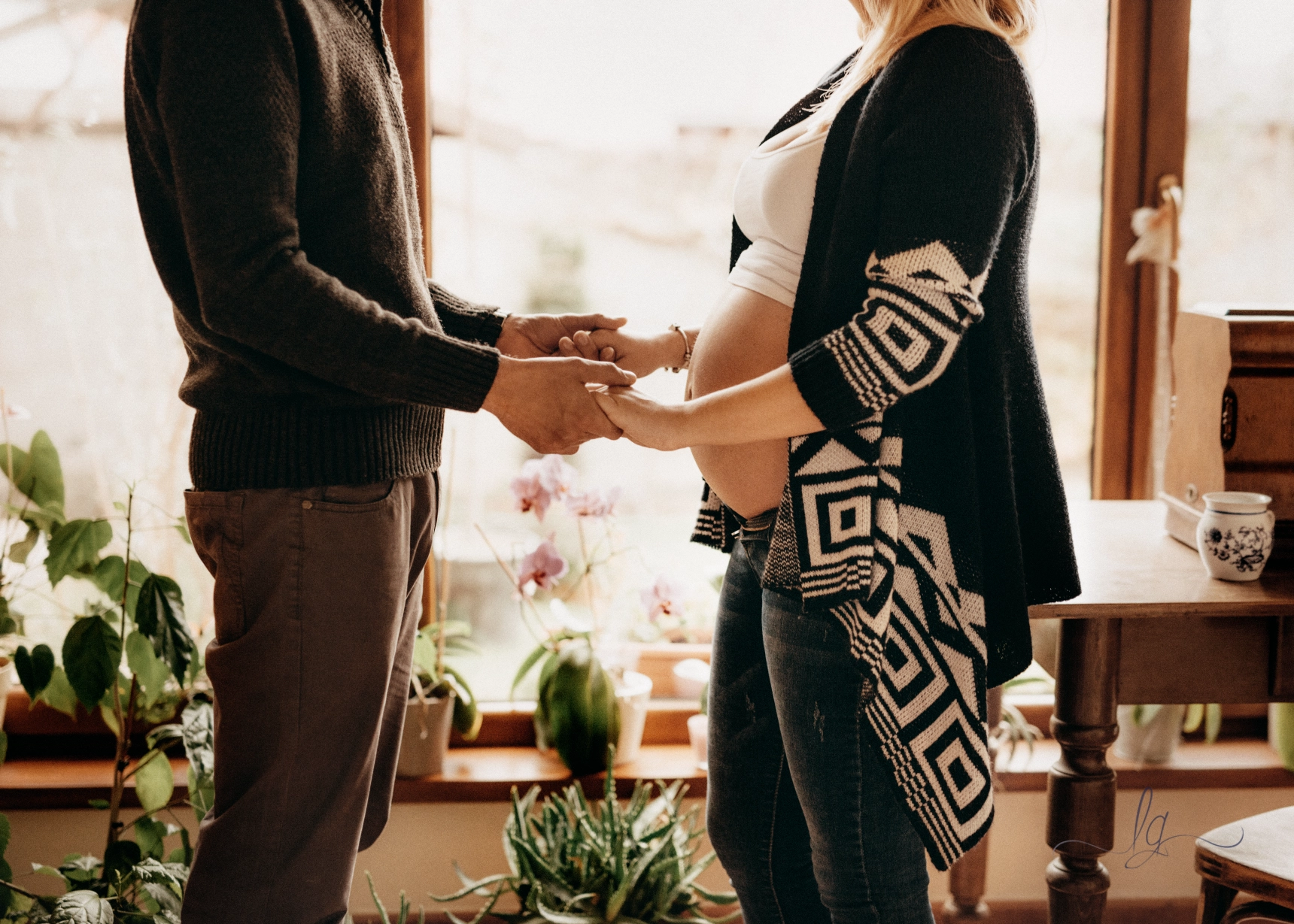 A maternity portrait of an expecting parents holding hands, baby bump visible by a sunlit window with potted plants and orchids.