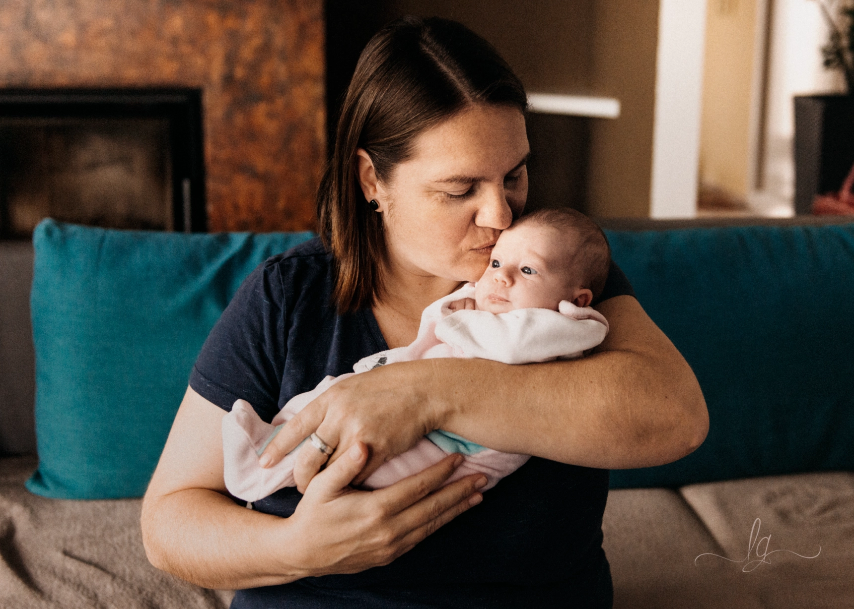A mother gently holding and kissing her newborn baby on the forehead and sitting on a green couch.