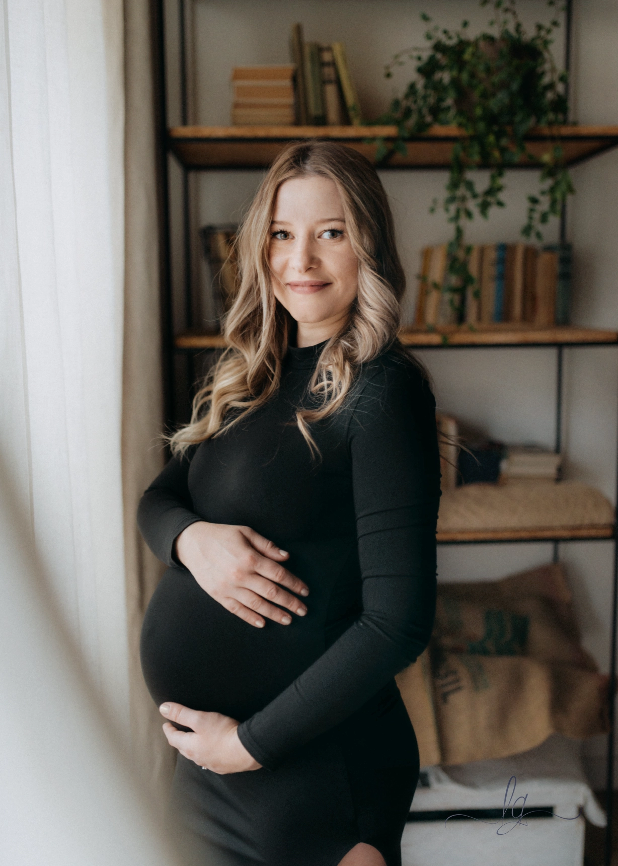 Portrét usmívající se těhotné ženy v přiléhavých černých šatech stojící u okna před knihovnou.
